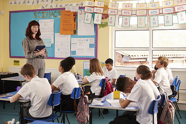 A teacher at the front of a classroom, talking to 8 students that are seated at a desk.